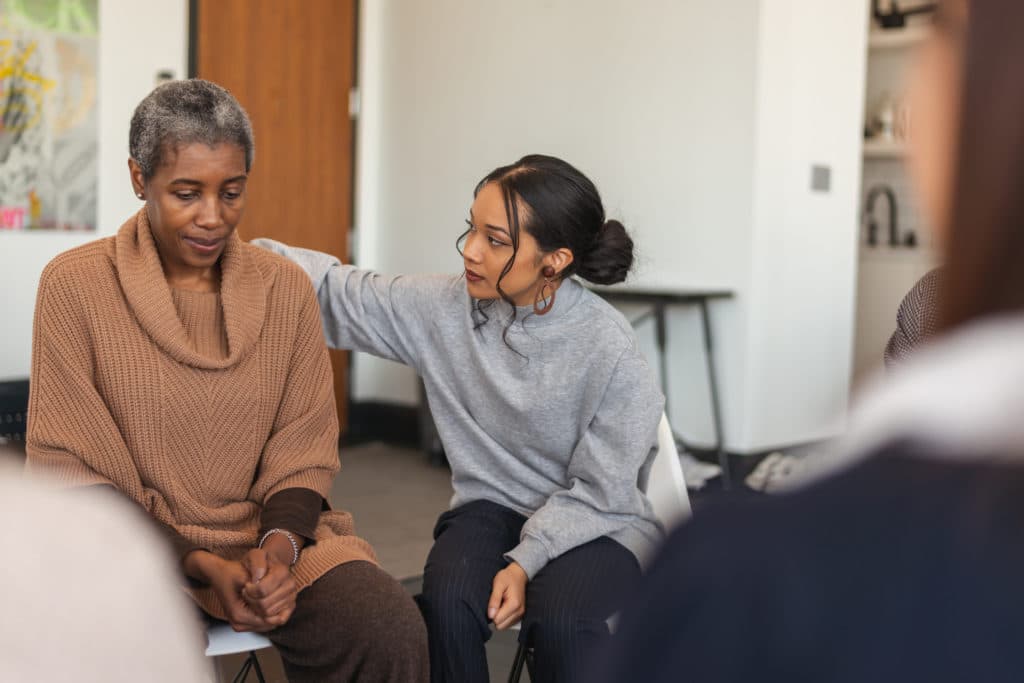 Young woman wearing a greay sweater comforts older woman wearing brown sweater by putting her arm round her shoulders in what appears to be a group setting.